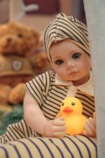 Reborn panenka Bonnie s realistickou silikonovou kůží, oblečená v pruhovaném oblečení, držící žlutou gumu.