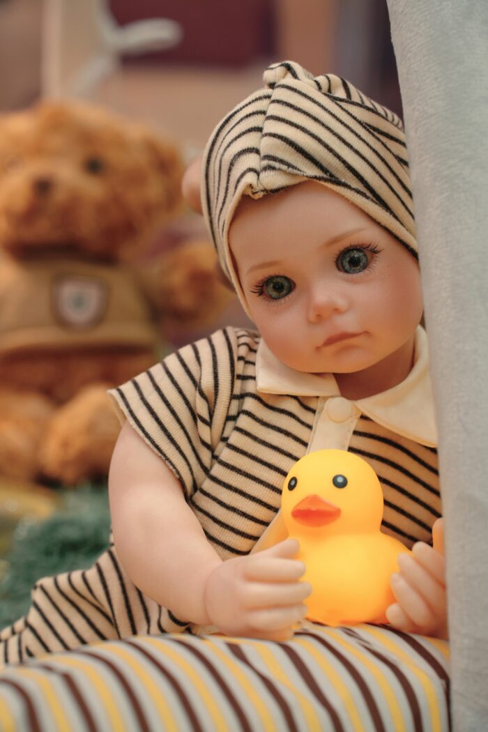 Reborn panenka Bonnie s realistickou silikonovou kůží, oblečená v pruhovaném oblečení, držící žlutou gumu.