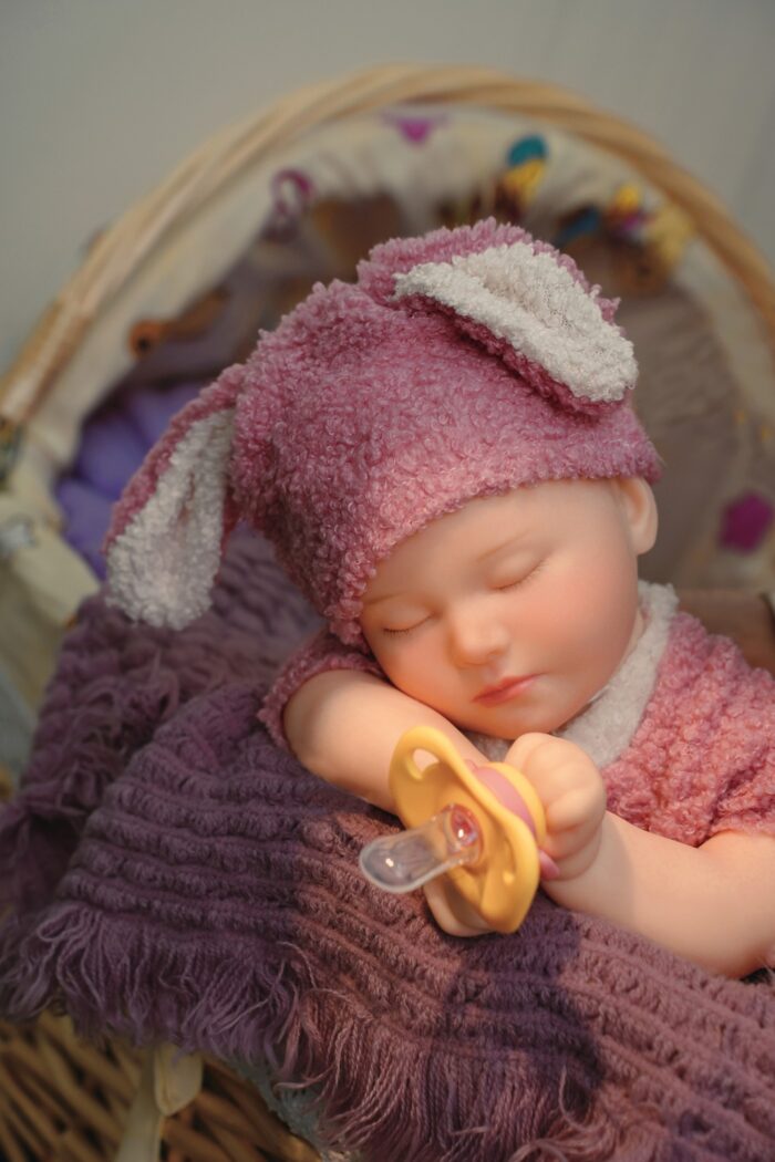 Realistic silicone reborn doll of a sleeping baby girl with a pink hat and pacifier.