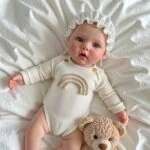 Realistic Kelly silicone reborn doll wearing a rainbow onesie and bonnet, holding a teddy bear.