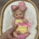 Atdzimusi lelle ar blondiem matiem, rozā bantīti un reālistiskām iezīmēm, ideāli piemērota kolekcionāriem un bērniem.