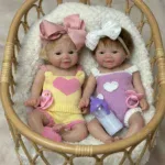 Reālistiskas silikona dvīņu lelles ar sakņotiem matiem, ģērbtas krāsainos tērpos, mājīgā pītā bundžā.