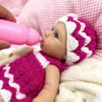 Silikonová panenka Reborn pije z růžové lahvičky, oblečená v růžovo-bílém pleteném oblečení.