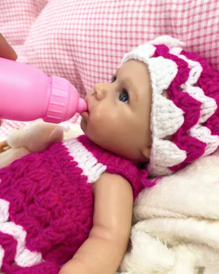 Silikonová panenka Reborn pije z růžové lahvičky, oblečená v růžovo-bílém pleteném oblečení.