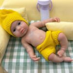 Poupée reborn réaliste en silicone avec des traits détaillés, un bonnet et un short jaunes en tricot, pour collectionneur.