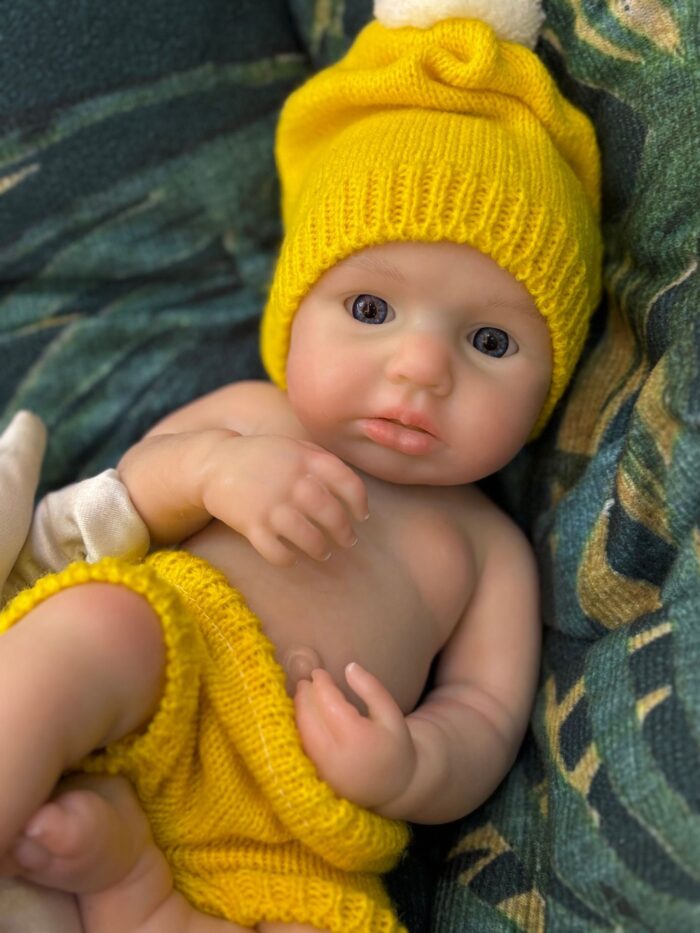 Poupée reborn en silicone, bébé portant un chapeau et une couche jaunes, présentant des traits réalistes et une peau douce.