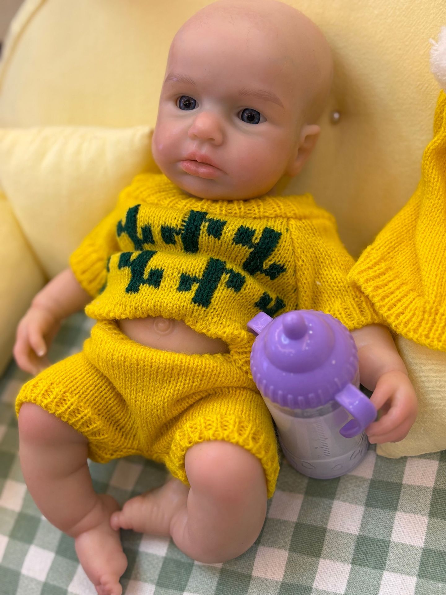 Poupée bébé reborn en silicone aux traits réalistes, vêtue de vêtements jaune vif, tenant un objet violet.
