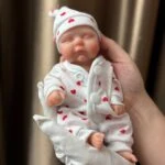 Reborn lutka koja nalikuje pravoj bebi, odjevena u bijelo s crvenim srcima, savršena za kolekcionare i entuzijaste.