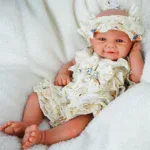 Reborn baby doll with detailed features, dressed in a ruffled cream outfit and matching hat, lying o.