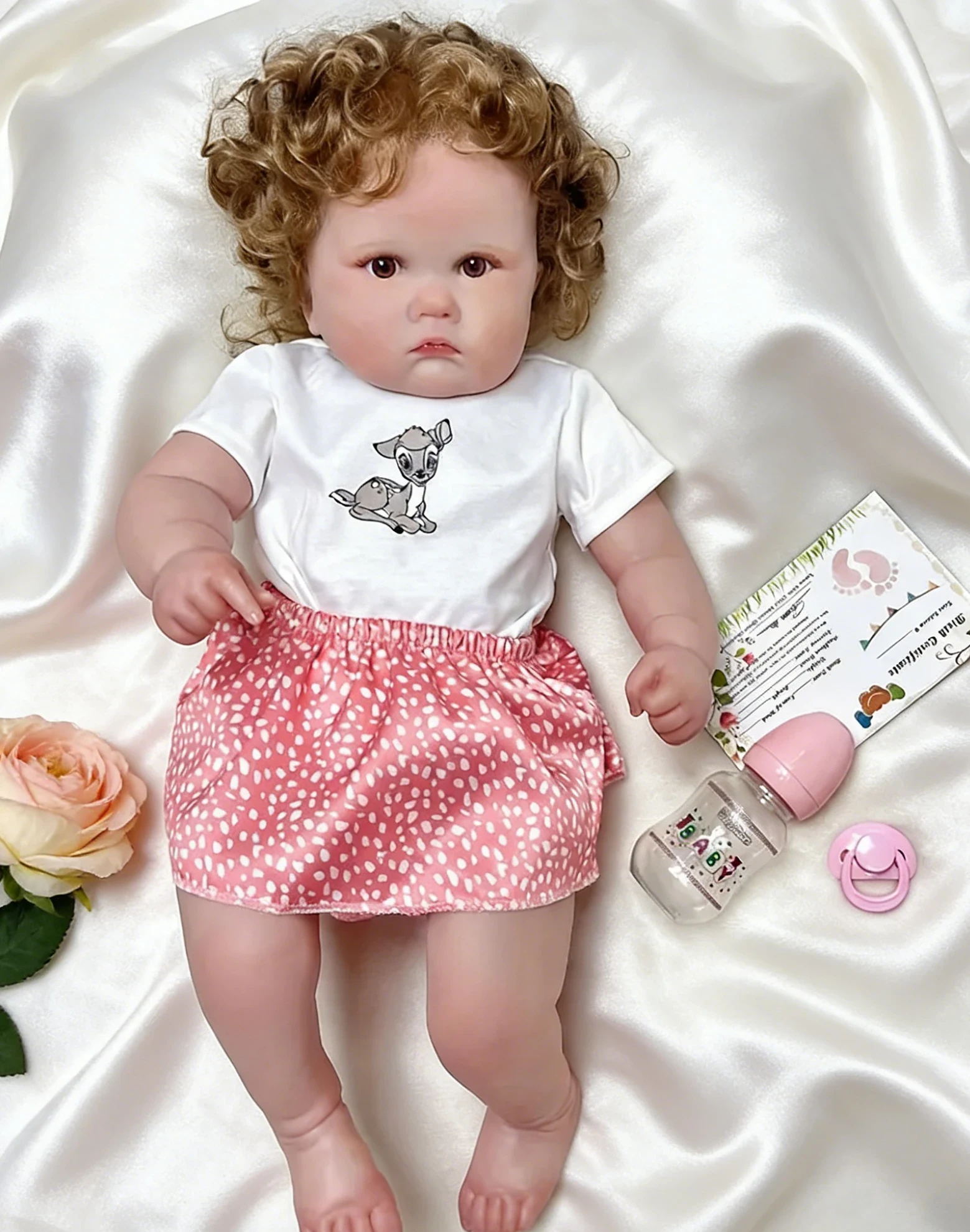 Irina The Real Reborn Dolls Silikoninė Vinilo Mergaitė 24 Inch 5 Realistiška atgimusi lėlė su garbanotais plaukais, apsirengusi miela balta palaidine ir rožiniu taškuotu sijonu.