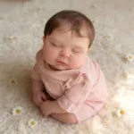 Realistic baby reborn doll sleeping on soft surface with daisies, lifelike details.
