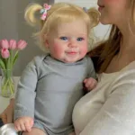 Yndig smilende babypige med blond hår og blå øjne, der holder en dukke, i hyggelige omgivelser.