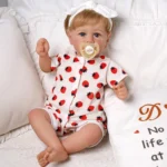 Poupée bébé réaliste aux yeux bleus et aux cheveux blonds, vêtue d'une tenue à imprimé fraise.