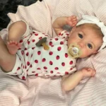 Poupée bébé réaliste aux yeux bleus, avec une tétine et une tenue à fraises, sur une literie rose.
