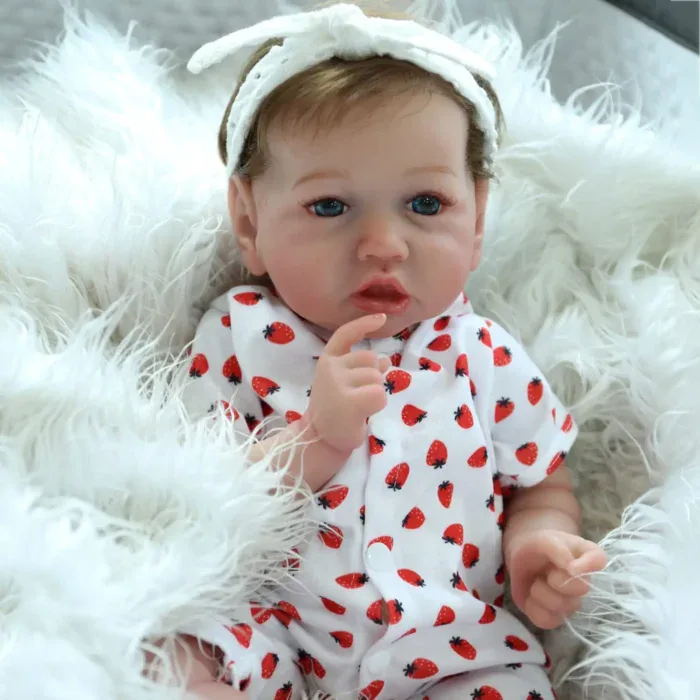 Poupée bébé réaliste aux yeux bleus et vêtue d'une robe à fraises, posée sur une couverture blanche moelleuse.