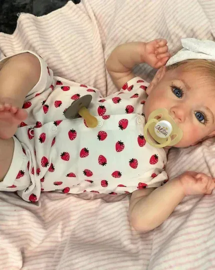 Poupée bébé réaliste aux yeux bleus, avec une tétine et une tenue à fraises, sur une literie rose.