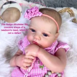 Realistická reborn bábika dievčatka v ružových kockovaných šatách a čelenke.