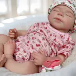 Păpușă fetiță adormită, îmbrăcată în haine roz cu motive florale și pălărie, piele realistă din silicon.