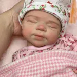 Păpușă reborn realistă, fetiță adormită, cu căciulă florală și ținută roz.
