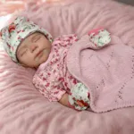 Păpușă adorabilă care adorme, cu căciulă cu motive florale și pătură roz.