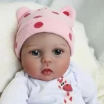 Realistische Babypuppe mit rosa gepunktetem Hütchen und weißem Outfit.