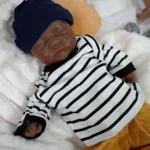 Poupée reborn réaliste de bébé garçon noir de Zion, vêtue d'une chemise rayée et d'un chapeau bleu marine.