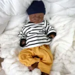 Poupée reborn réaliste, bébé à la peau foncée, vêtue d'une chemise rayée et d'un pantalon jaune.