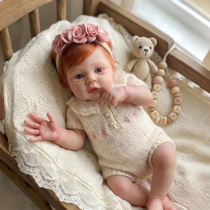 Realistic red-haired baby girl doll with pink headband in crib.