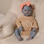 Blue-skinned girl reborn doll with yellow eyes and pink bow, sitting on cream blanket.