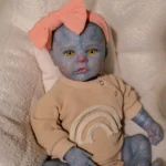 Blue-skinned reborn doll with yellow eyes, pink bow, and rainbow sweater.