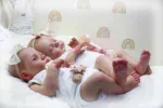 Zwei Zwillings-Reborn-Babypuppen schlafen auf einem Bett mit Regenbogentapete.