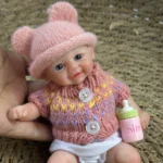 Niedliche Babypuppe mit rosa Mütze, Strickpullover und Milchflasche vor gewebtem Hintergrund.