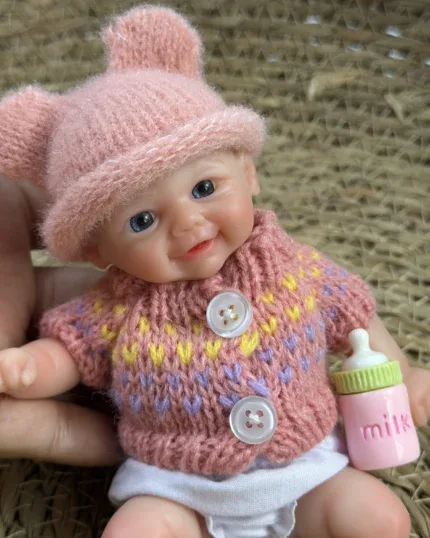 Niedliche Babypuppe mit rosa Mütze, Strickpullover und Milchflasche vor gewebtem Hintergrund.
