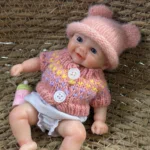 Entzückende, pummelige Reborn-Babypuppe mit rosa Mütze und Pullover, sitzend auf einer gewebten Oberfläche.